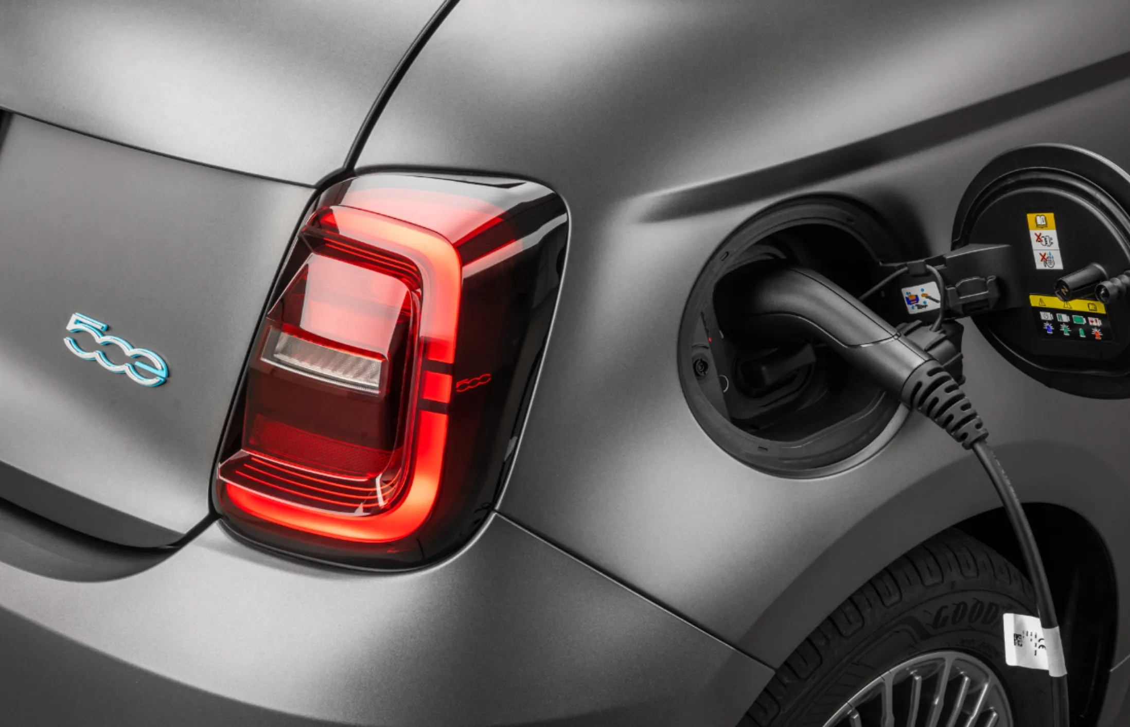 Lateral traseira do Fiat 500 elétrico, na cor prata, com carregador elétrico conectado ao carro. 